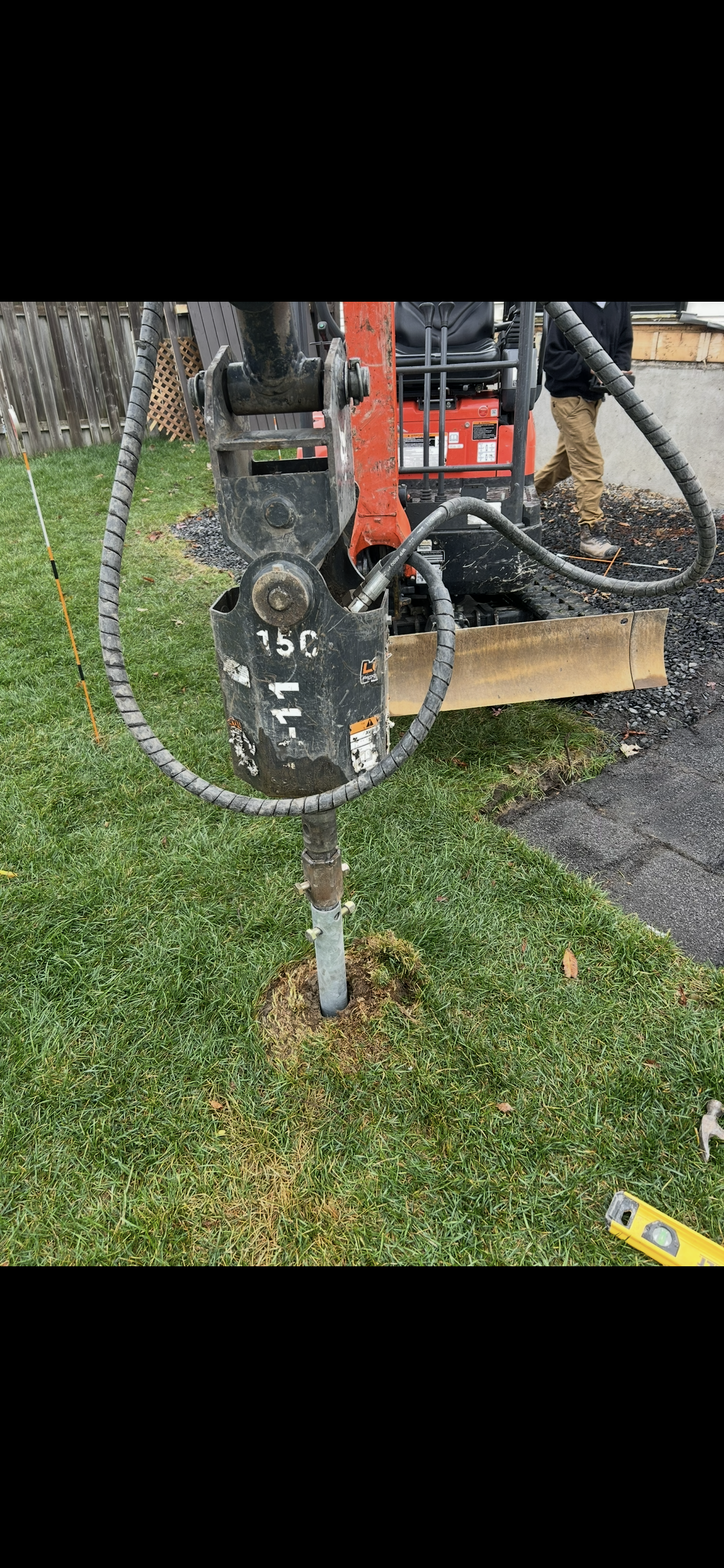 Une mini-pelle orange équipée d'un marteau hydraulique enfonce un poteau métallique dans une pelouse.