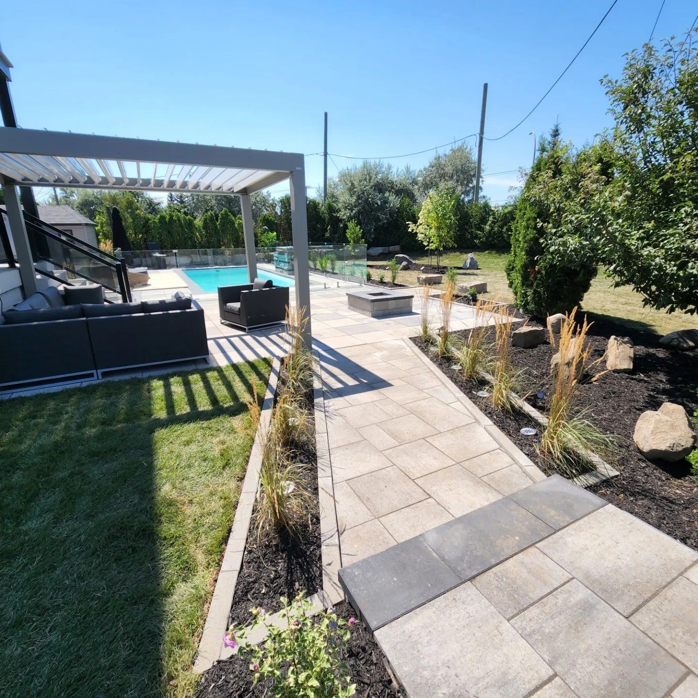 Une terrasse ensoleillée à l'arrière comprend une pergola au-dessus d'un coin salon extérieur, une allée pavée, un foyer et une piscine creusée.