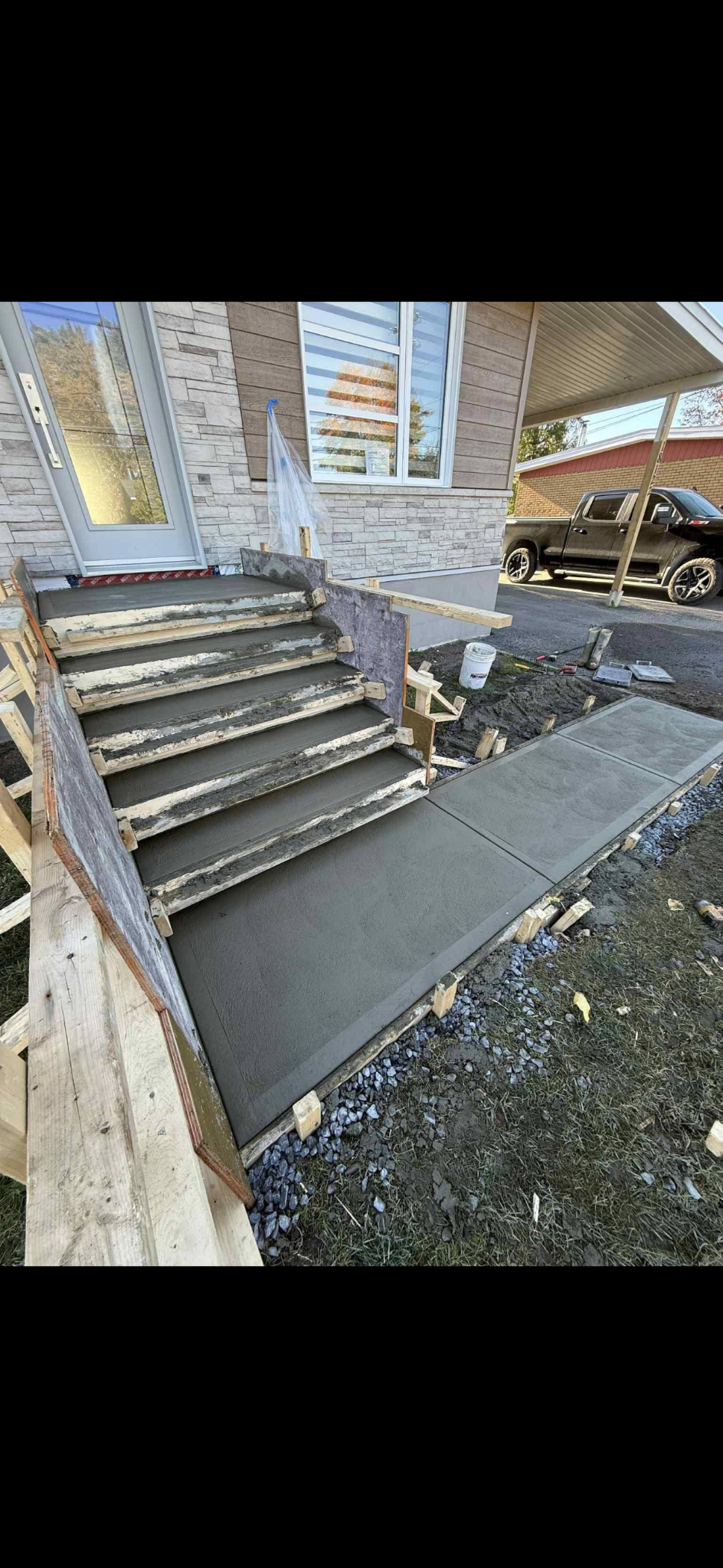Escalier en béton fraîchement coulé et allée menant à l'entrée d'une maison d'habitation, encadrés par des coffrages de construction en bois.