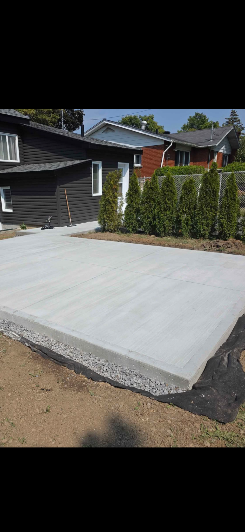 Une dalle de patio en béton fraîchement coulée à côté d'une maison noire, avec une rangée de conifères en arrière-plan.