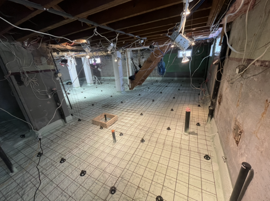 Un sous-sol non aménagé avec un plancher en treillis métallique posé sur une surface en béton, des tuyaux de plomberie et des solives de plafond apparentes.