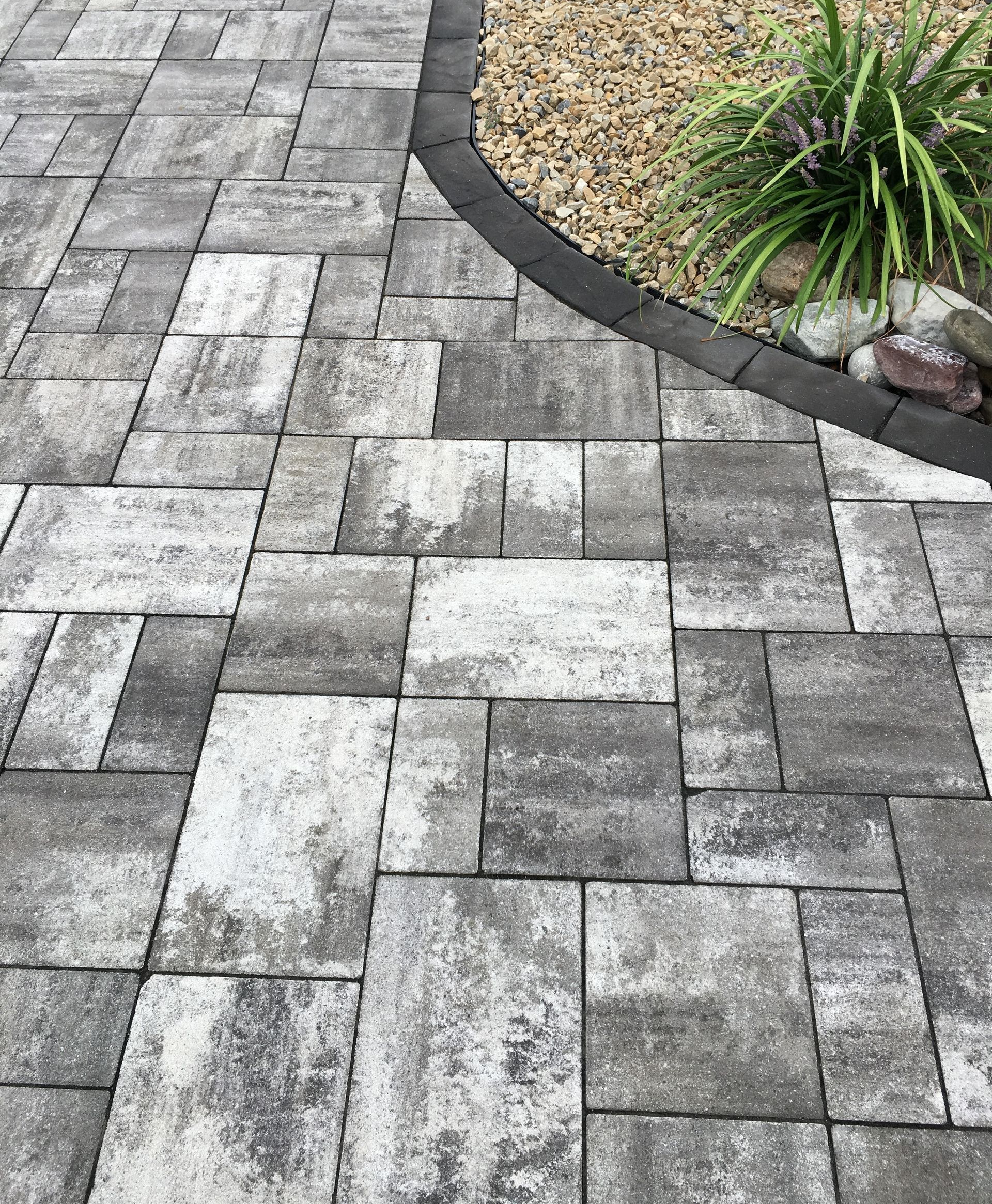 Des pavés rectangulaires gris disposés selon un motif géométrique, bordés de pierres sombres incurvées, à côté de gravier et d'une plante.
