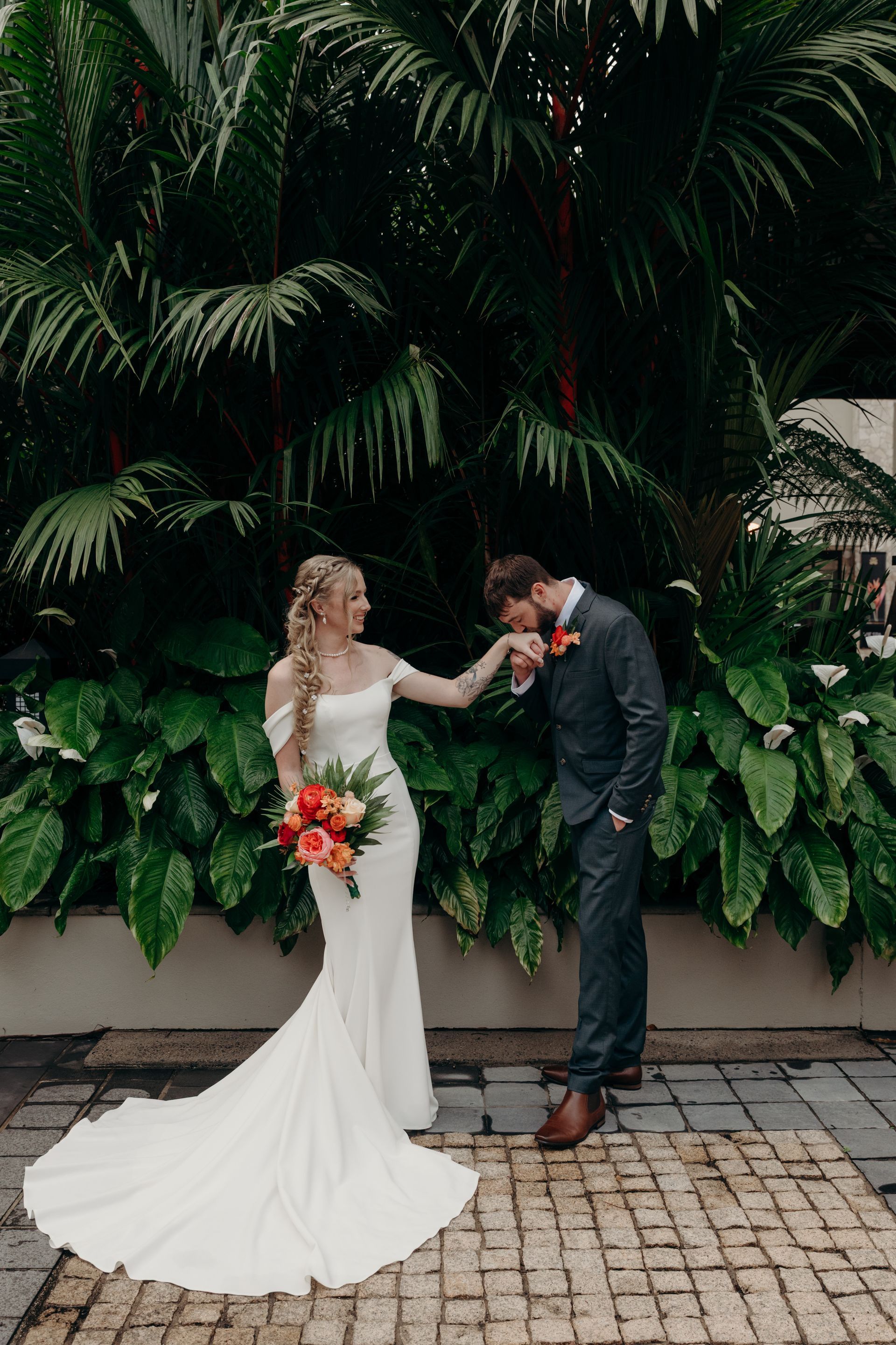 Bride in white gown holding bouquet, groom kissing her hand in front of lush green foliage - GinaS Cairns Tropical Weddings in Parramatta Park, QLD