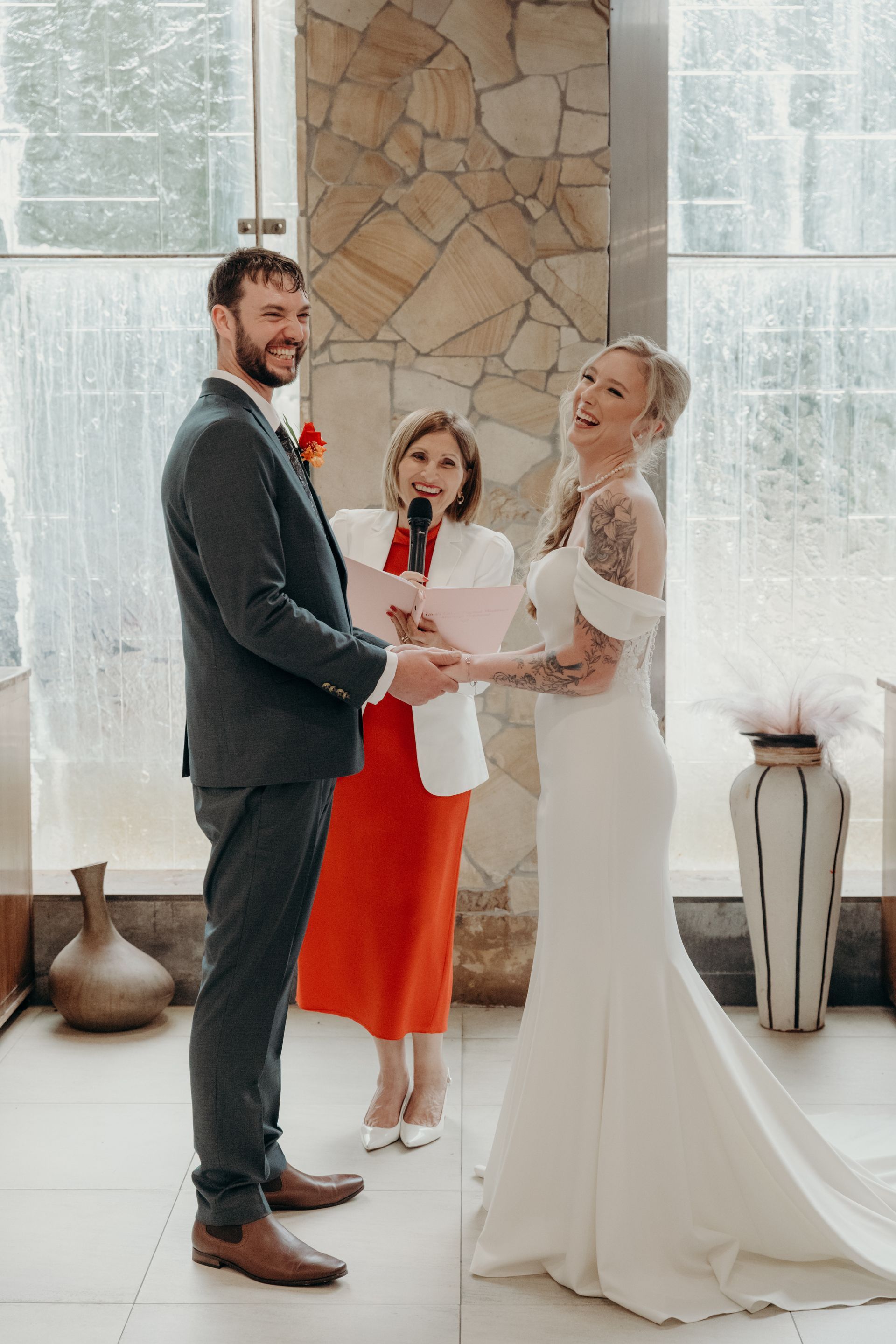 Couple holding hands during wedding ceremony, laughing. Officiant in red dress. Indoor setting with large window - GinaS Cairns Tropical Weddings in Parramatta Park, QLD