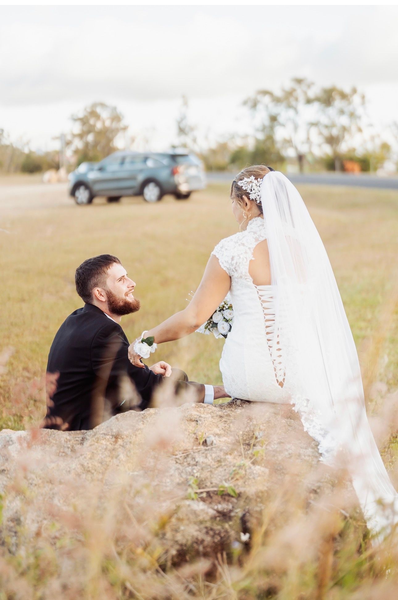 Bride in white dress and veil stands before a kneeling man in a field, with a car in the background.