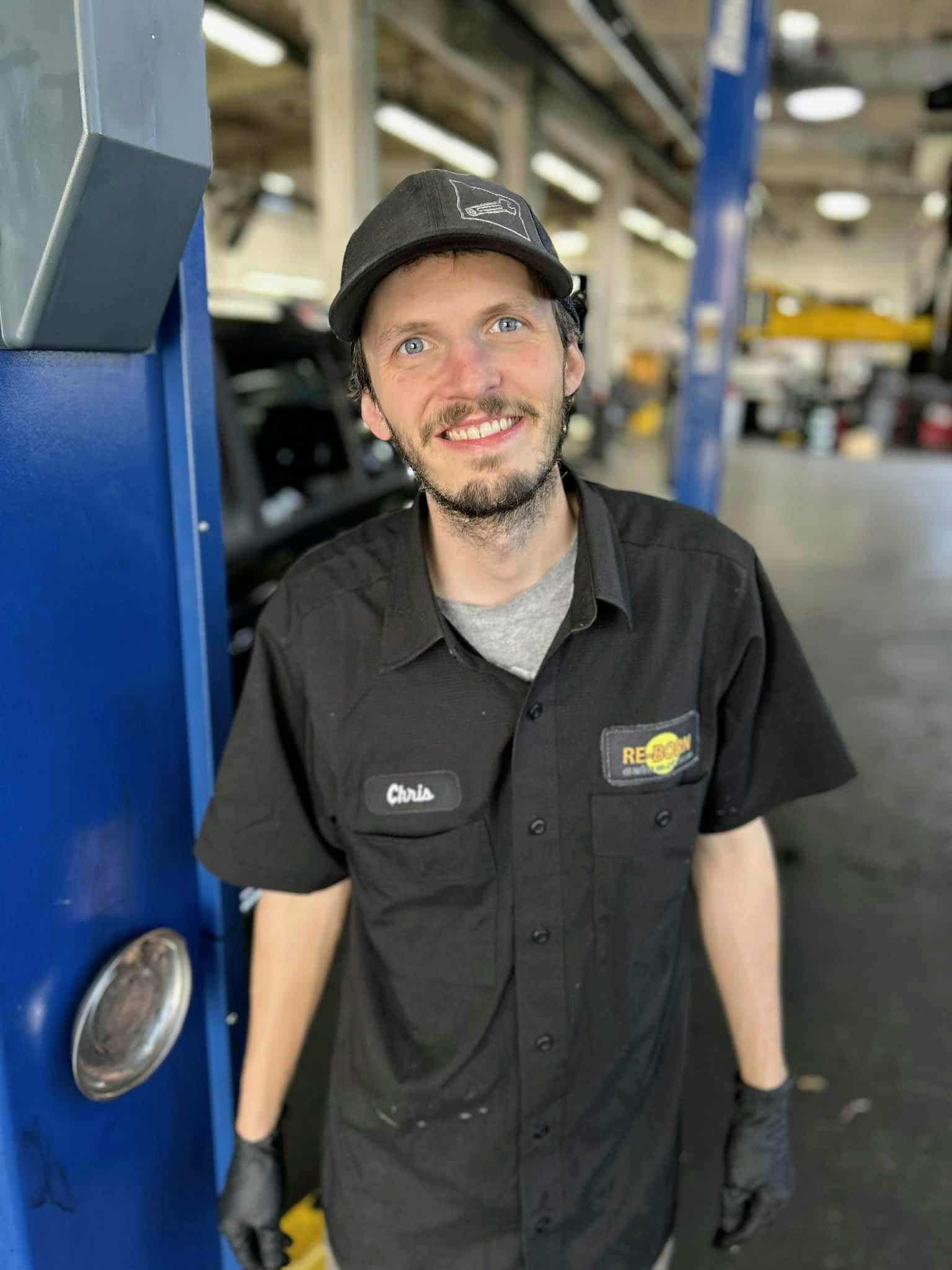Mechanic in black uniform smiles in a garage, wearing a hat and gloves.