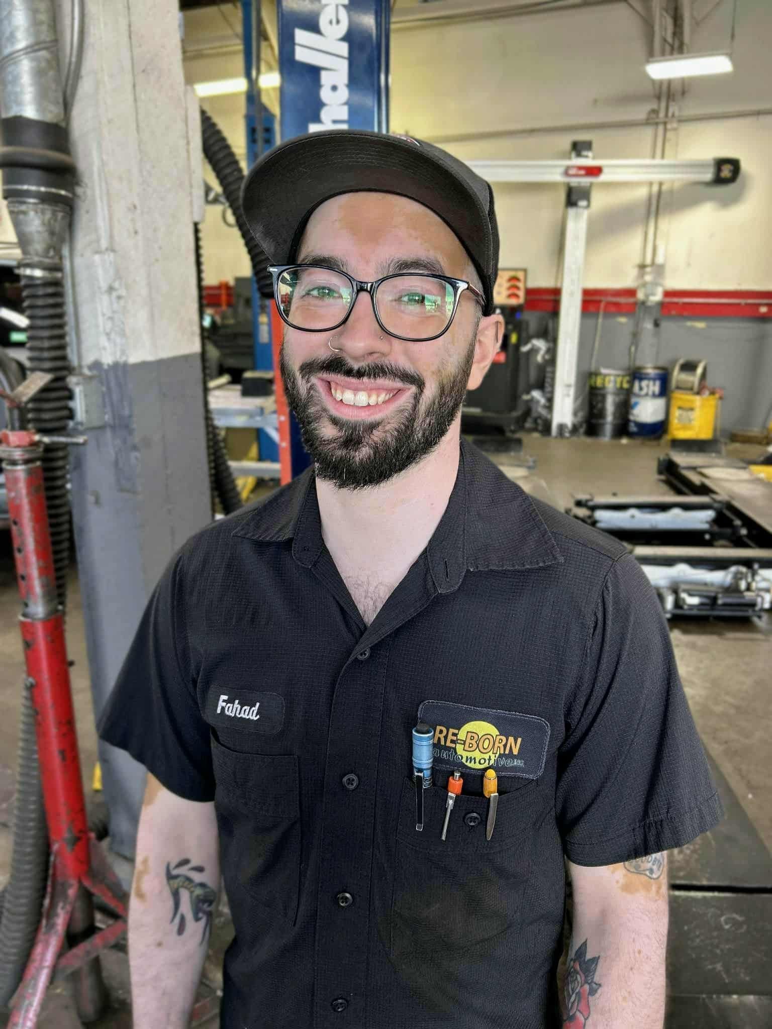 Mechanic smiling in a shop, wearing a black cap, glasses, and uniform. He has tattoos on his arms.