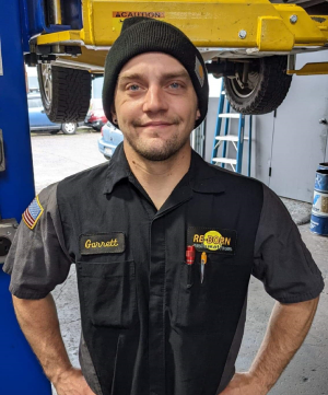 Mechanic Garrett in a shop, wearing a black cap and uniform, smiling, with hands on hips.