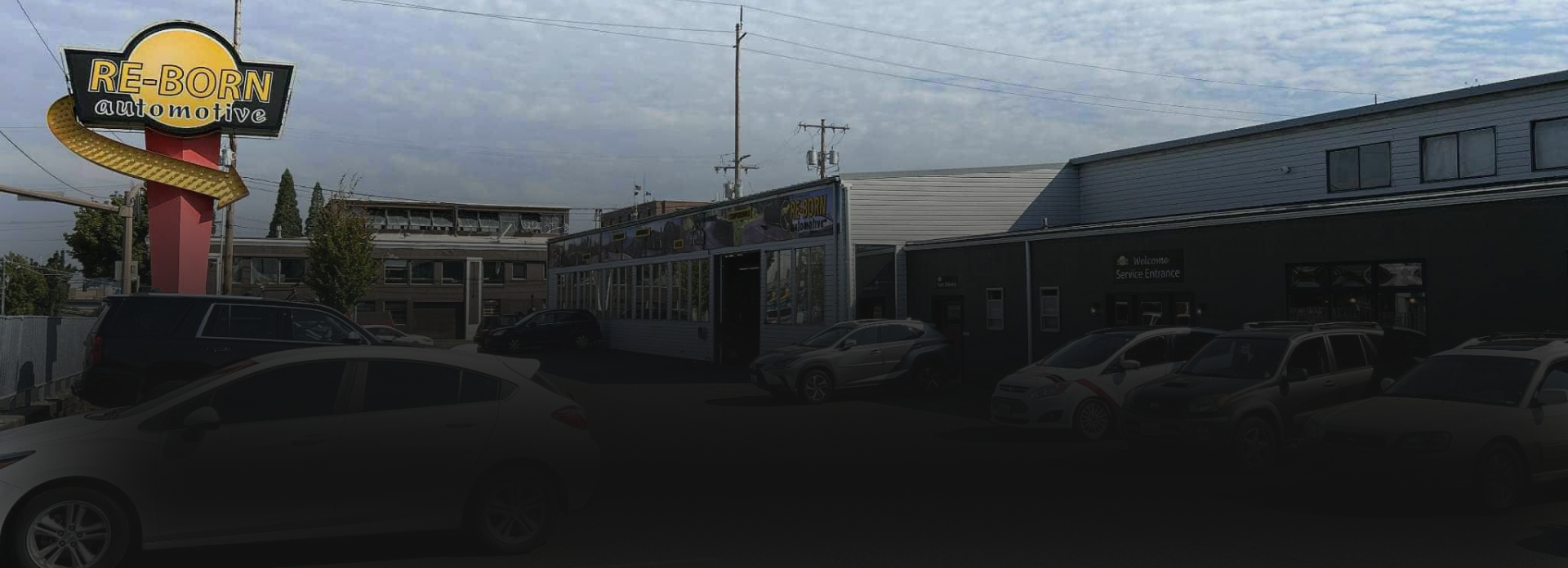 A group of people posing for a photo in front of a business, mostly men in dark shirts, with cars visible in the background. | Re-Born Automotive, Inc.