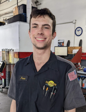 Mechanic smiling in a garage, wearing a work uniform with tools in his pocket and an American flag patch.