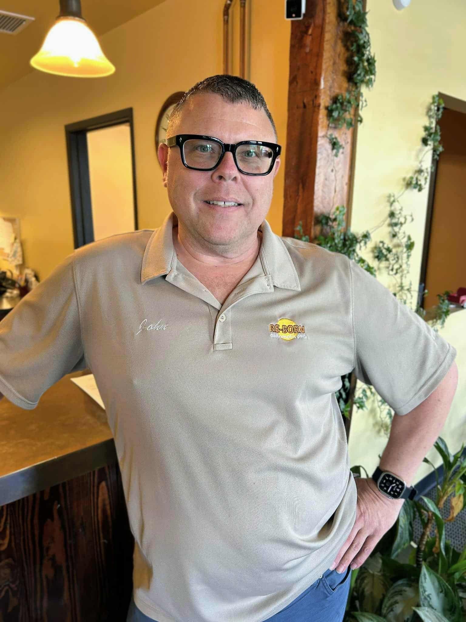 Man with glasses, wearing a tan polo shirt, smiling, hands on hips, indoors.