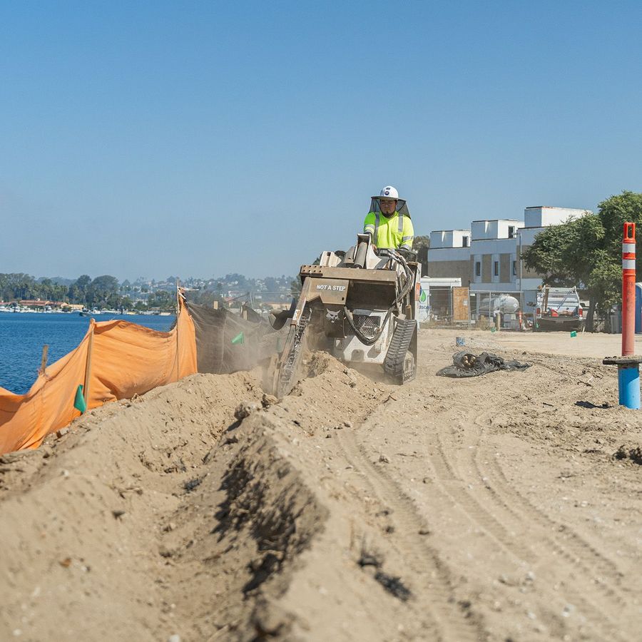 Construction worker operating a skid steer on a sandy beach near water, under a blue sky.