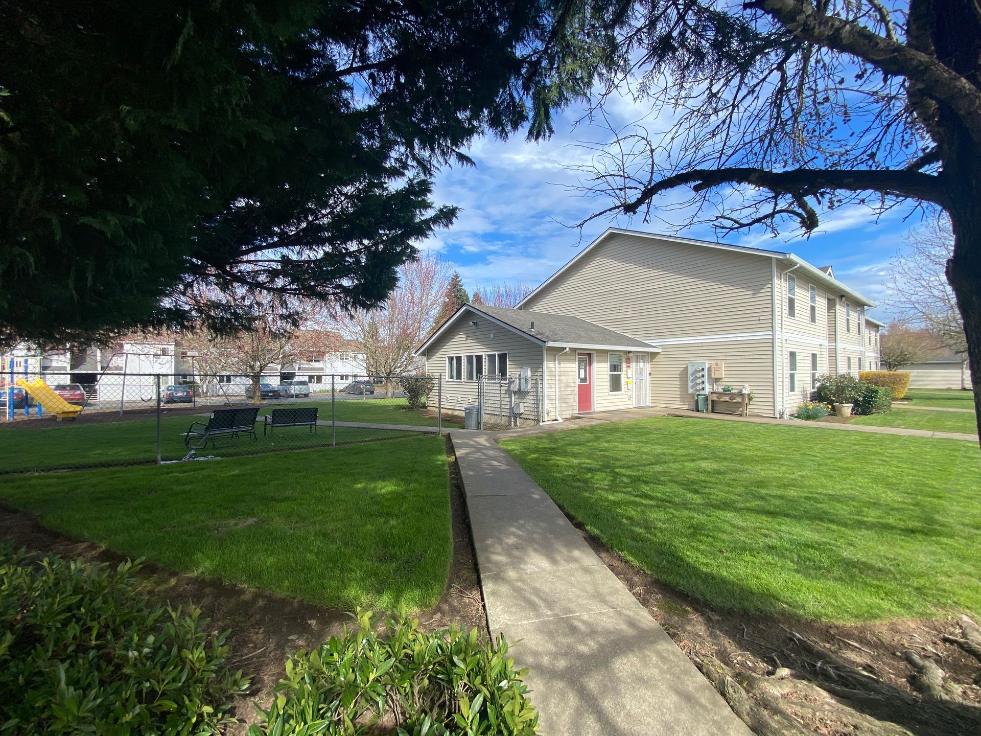 A concrete path leads through a grassy yard toward a two-story beige apartment building with a red door.