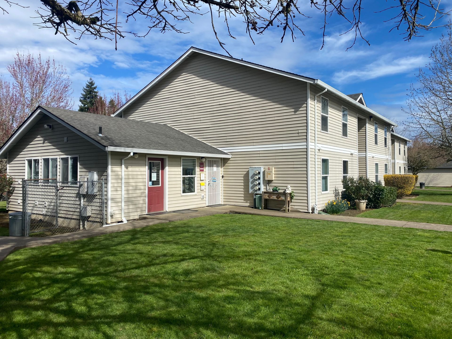 A beige two-story apartment building with a single-story entry, a red door, and a grassy front lawn under a blue sky.