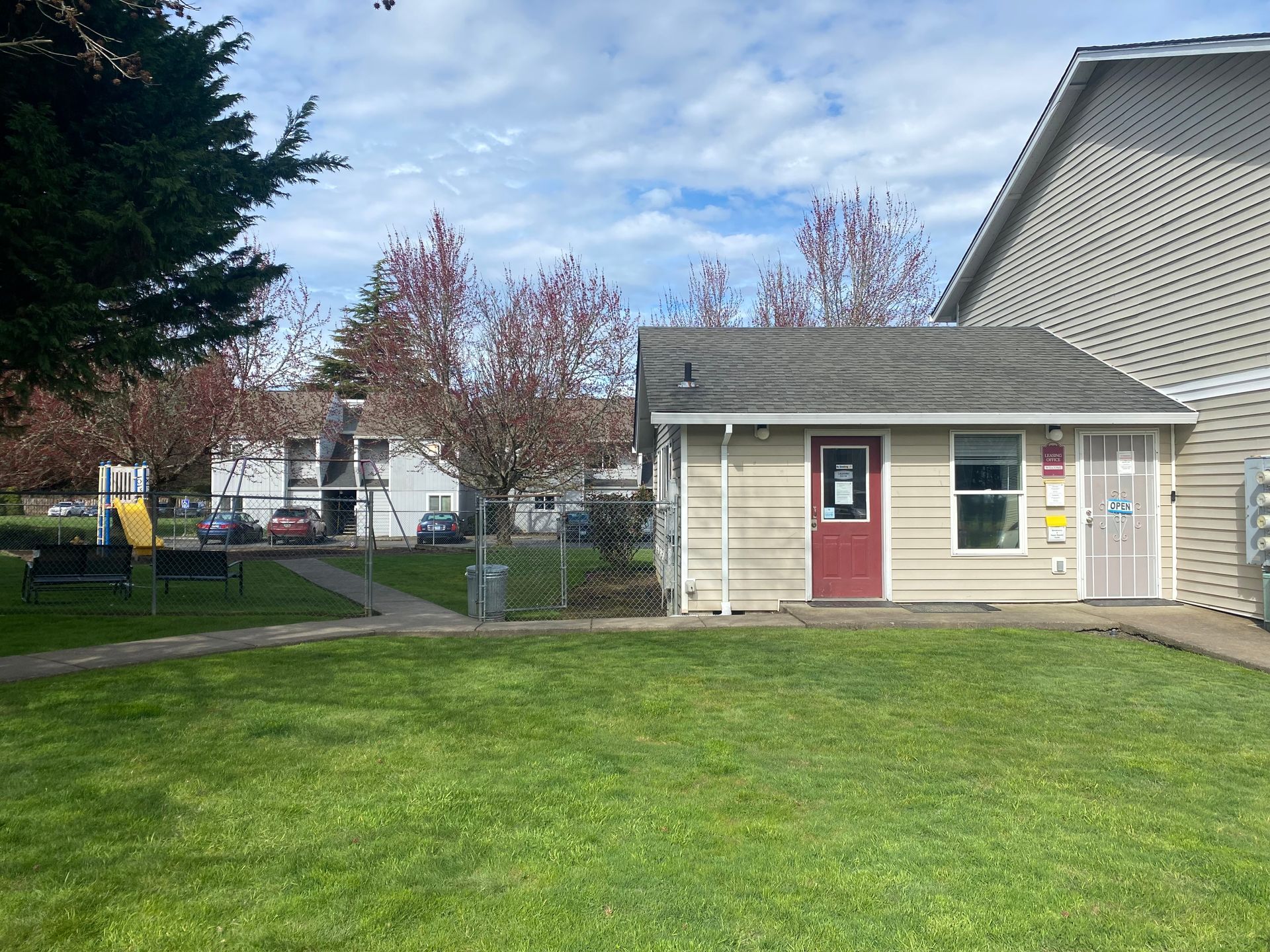 A single-story office building with a red door and tan siding, set next to a grassy lawn and a partially visible playground.