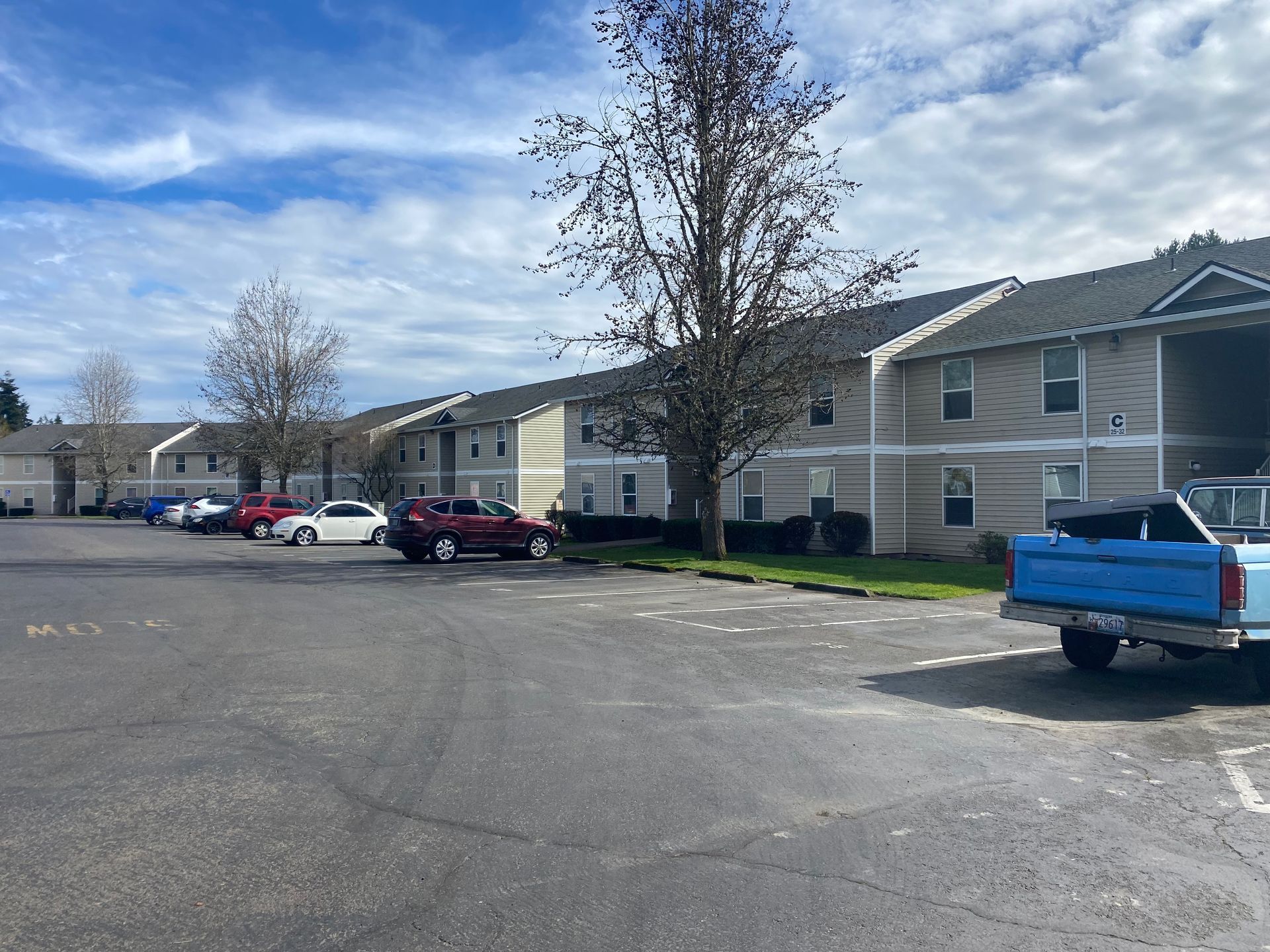 A multi-story beige apartment building with a parking lot containing several cars and a blue truck under a cloudy sky.
