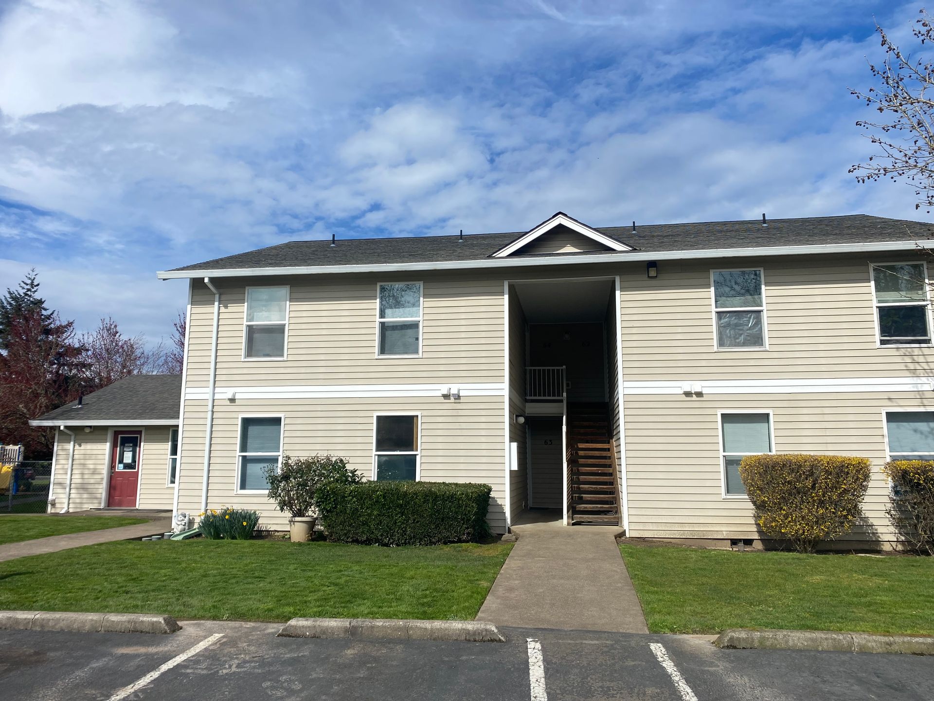 A two-story apartment complex with beige horizontal siding, a central stairwell, and a dark shingled roof under a blue sky.