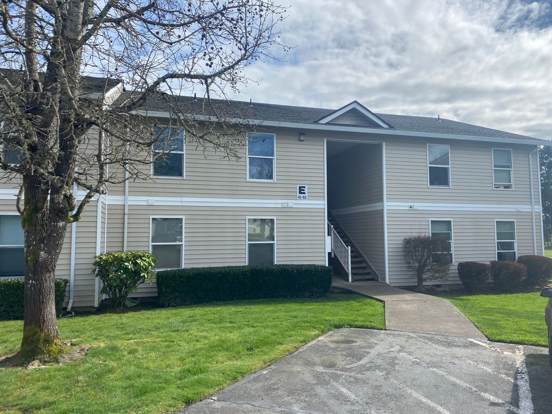 A two-story apartment building with beige siding, a dark roof, and an open stairwell entry under a cloudy sky.