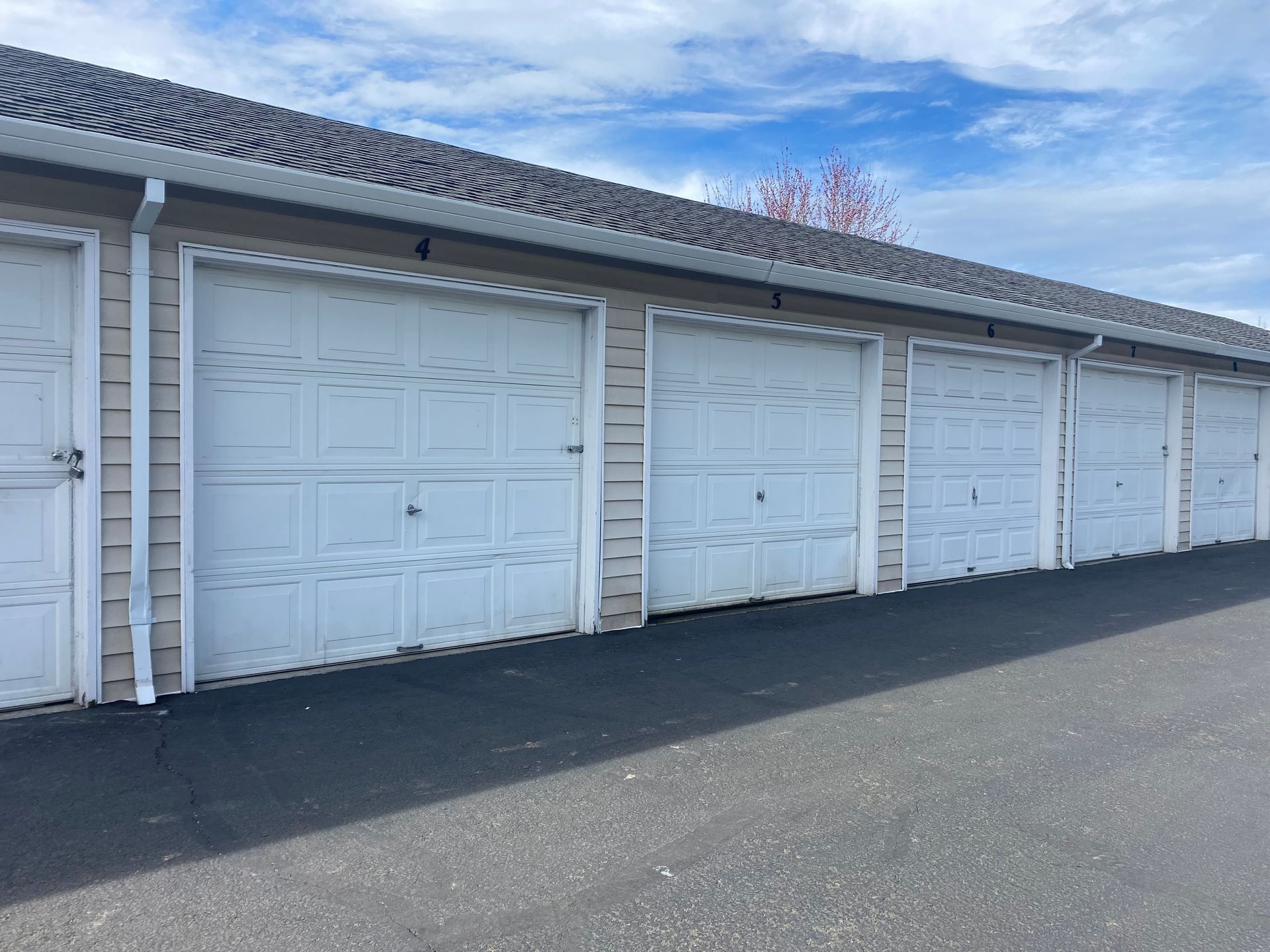 A row of white, paneled garage doors on a light-colored building with a gray asphalt driveway under a blue sky.