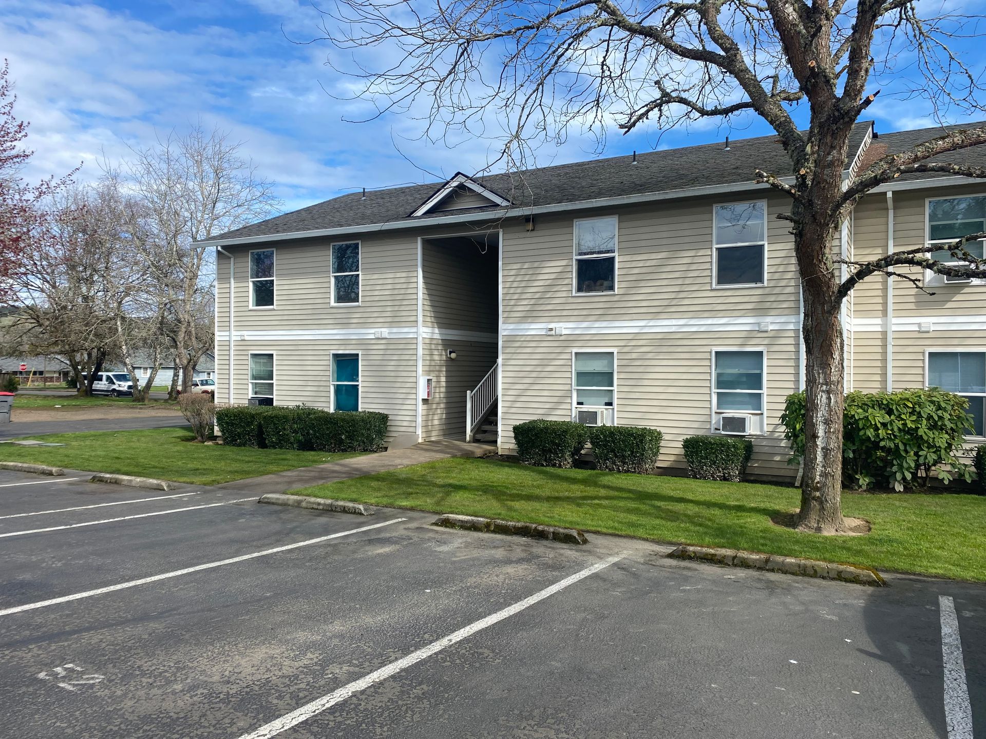 A two-story, light-colored apartment building with a central staircase, surrounded by lawn and an asphalt parking lot.