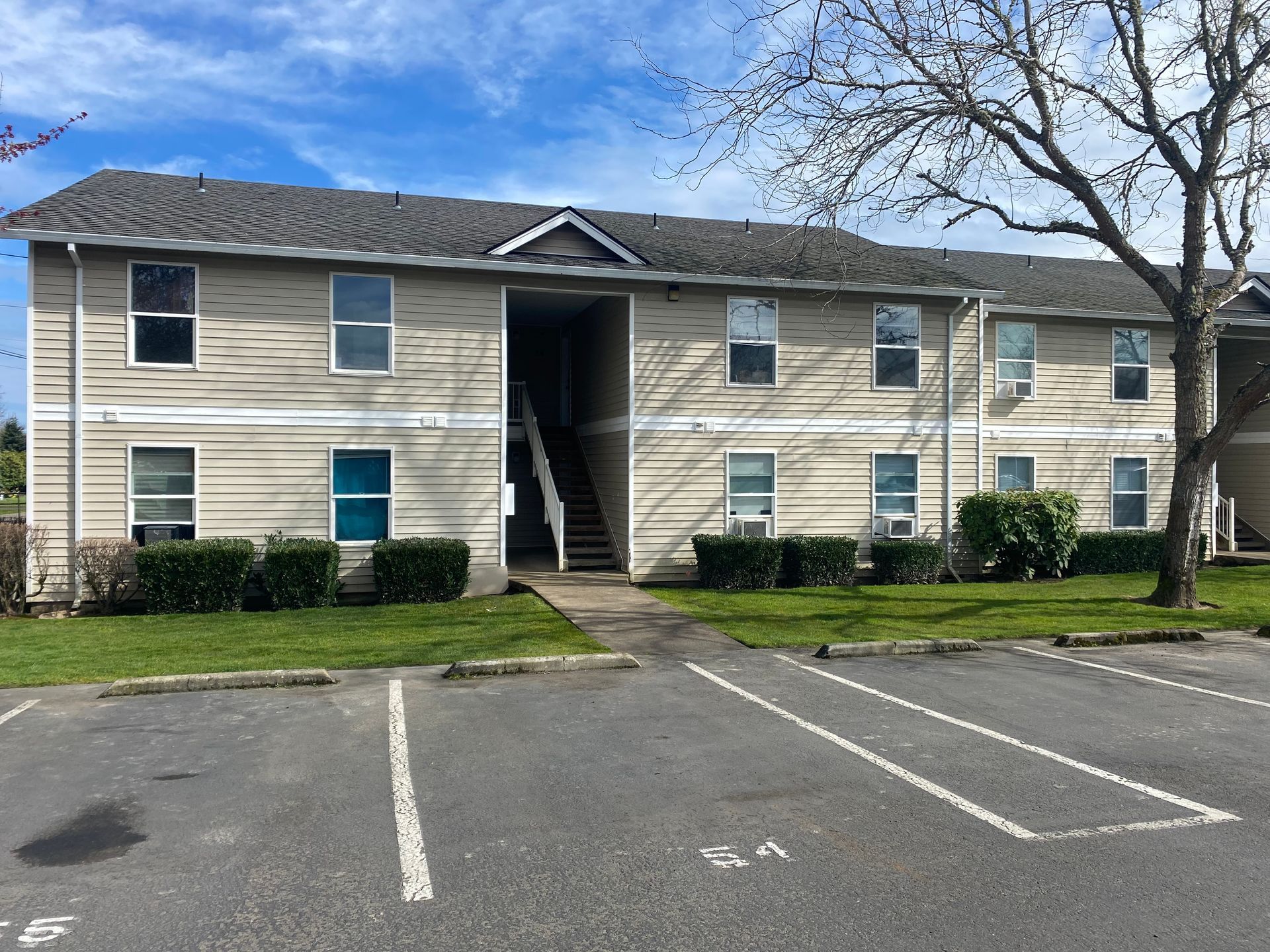 A two-story apartment complex with beige horizontal siding and a gray shingled roof, seen from a parking lot.