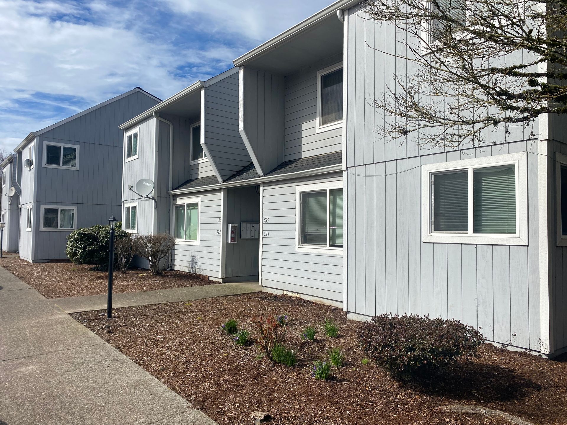 A row of two-story, gray-sided apartment buildings with white window frames, surrounded by wood-chip landscaping.
