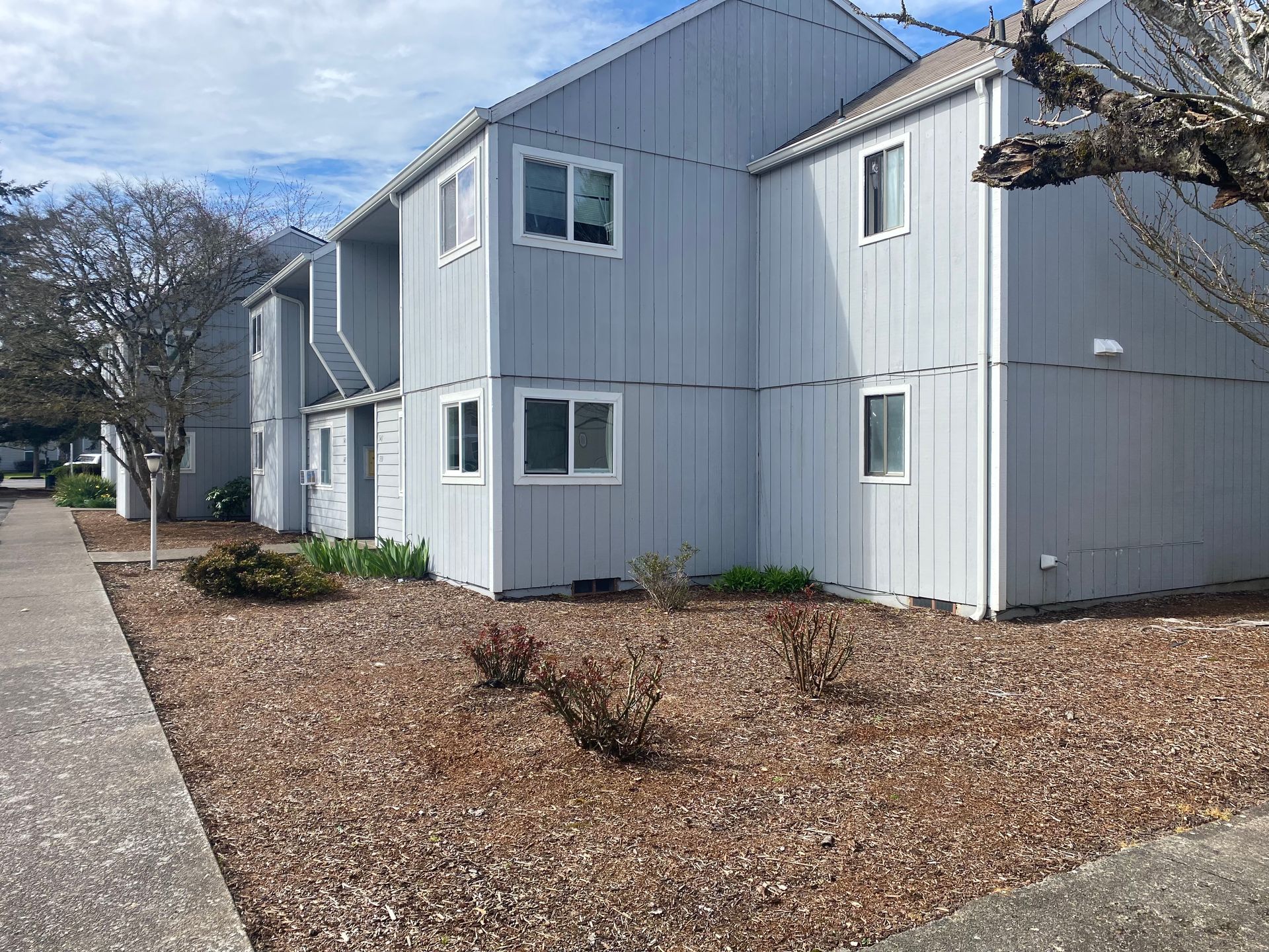 A two-story grey apartment building with a concrete sidewalk leading past a mulched yard with small shrubs.
