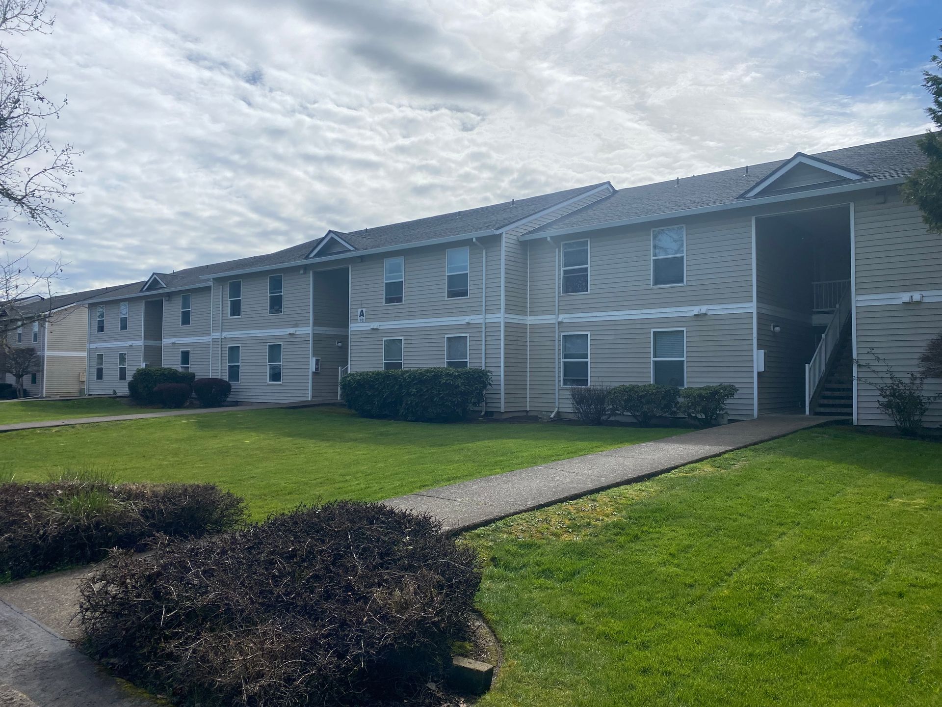 A two-story, beige apartment building with a gray roof, lawn, and a sidewalk leading to an open entrance.