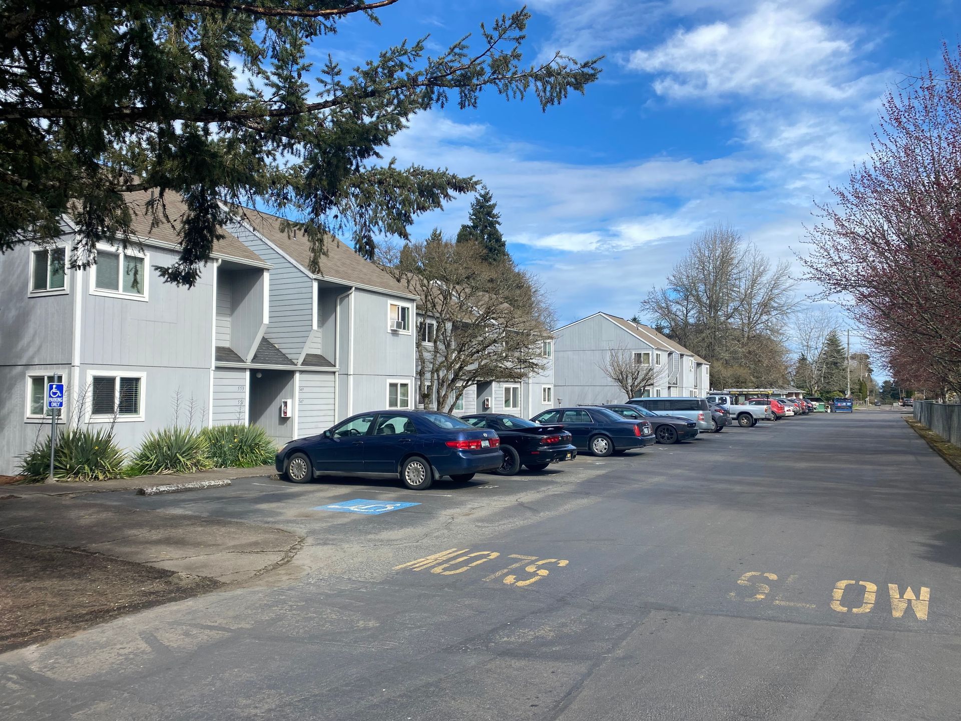 A parking lot in front of two-story grey apartment buildings under a blue sky, with a blue accessible parking sign.