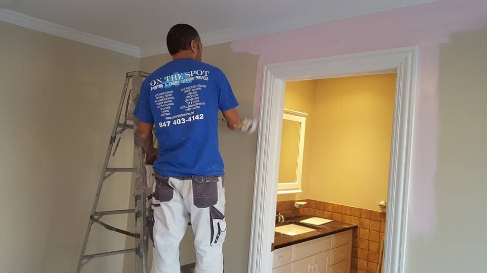 A man in a blue shirt is standing on a ladder painting a wall.