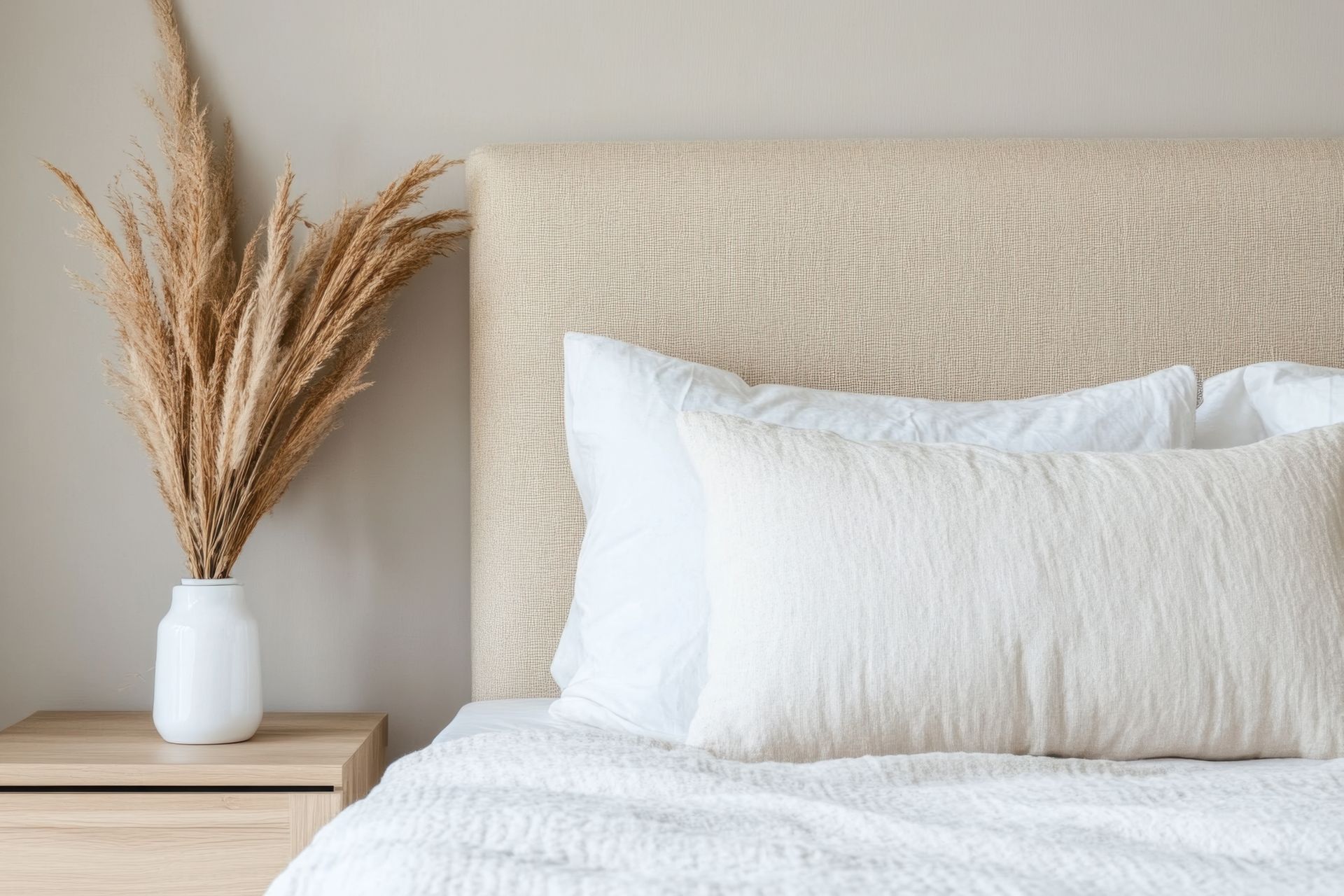 Bed with white linens, textured headboard, and bedside table with vase of dried flowers.