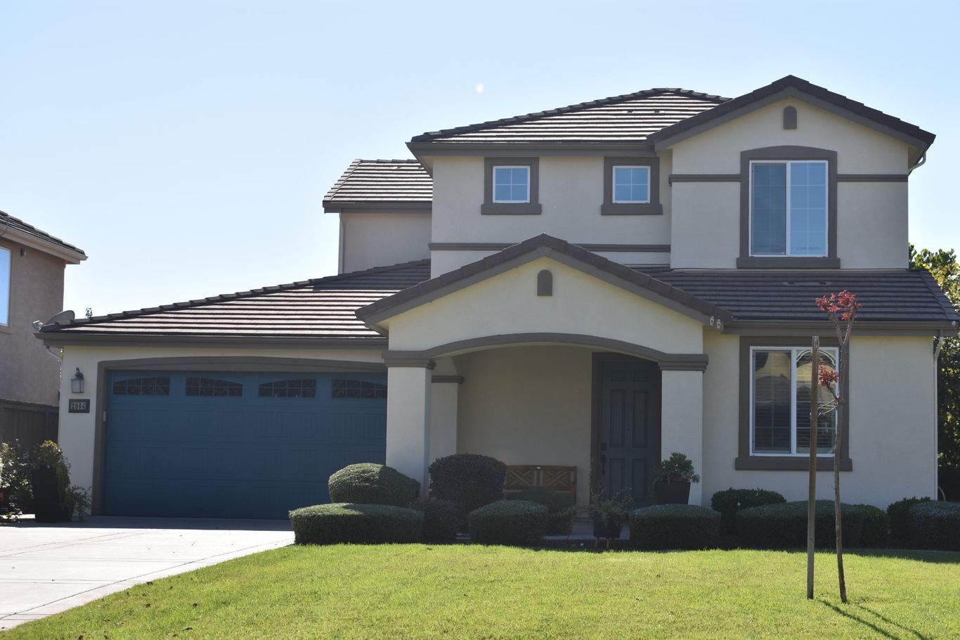 Two-story suburban house with a beige exterior, a dark tiled roof, a large blue garage door, and a manicured front lawn.