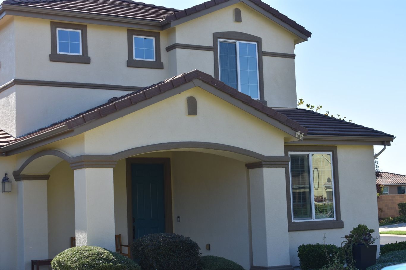 A beige two-story house with a brown tiled roof, a front porch, and a dark blue front door under a sunny blue sky.