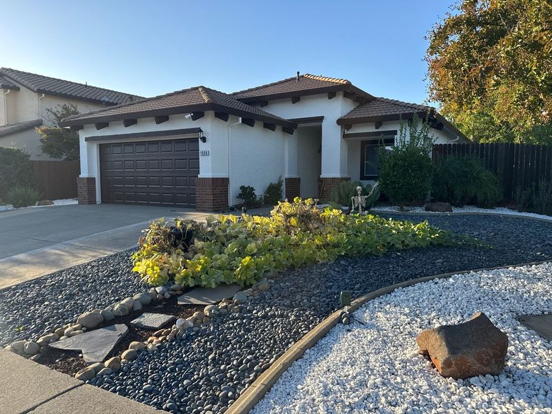 Single-story beige stucco house with a brown tile roof and garage, featuring a landscaped front yard with gravel beds.