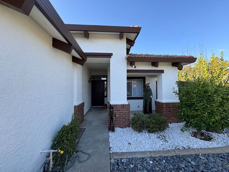 A tan stucco house with brown trim and brick accents features a recessed front entryway, white rock garden, and bushes.