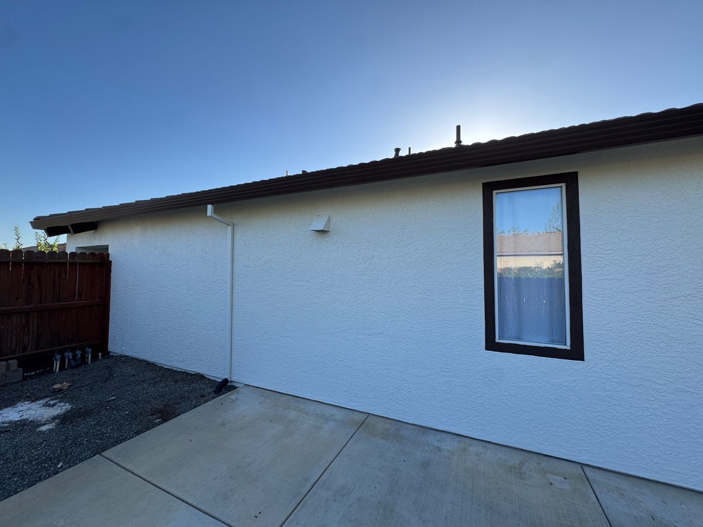 White textured exterior wall of a house with a window, a drain pipe, and a concrete patio under a blue sky.