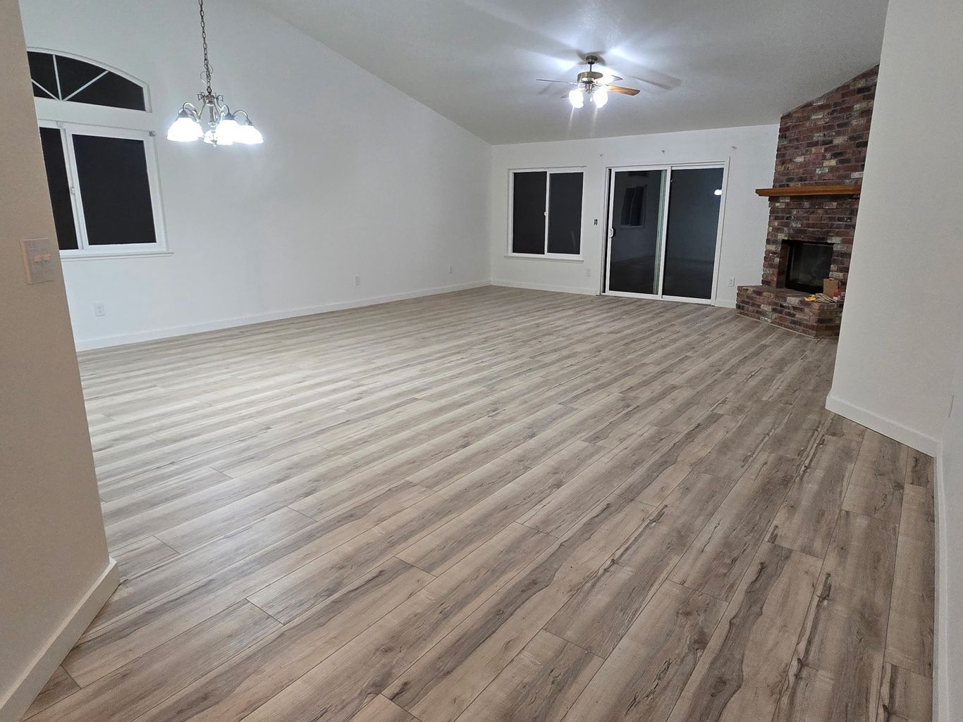 An empty, spacious living room with wood-look plank flooring, a vaulted ceiling, a chandelier, and a brick fireplace.