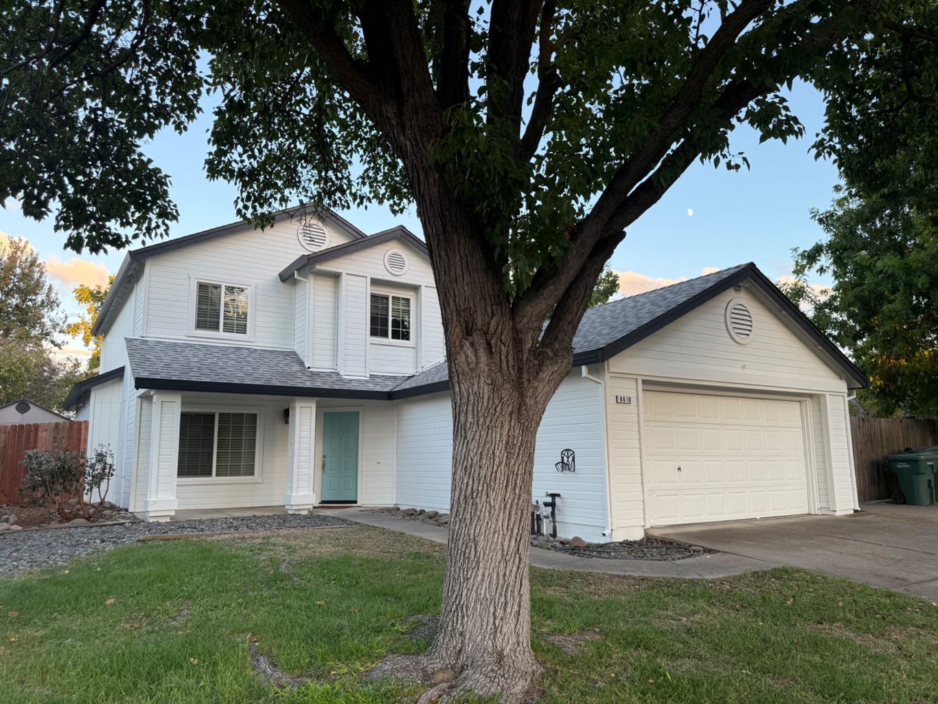 A two-story white house with a blue front door and an attached garage, partially obscured by a large tree in the front.
