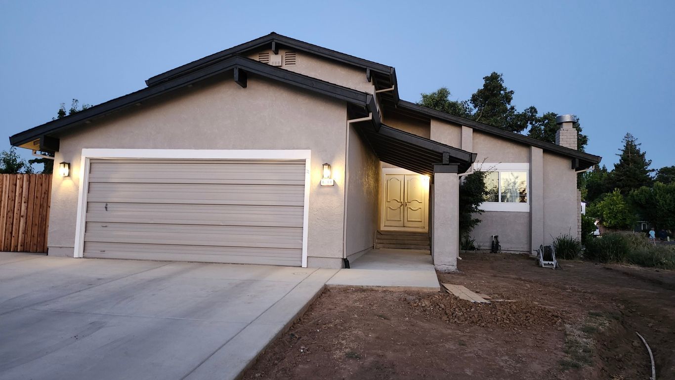 A single-story suburban house with a tan stucco exterior, a dark tiled roof, and a garage at twilight.