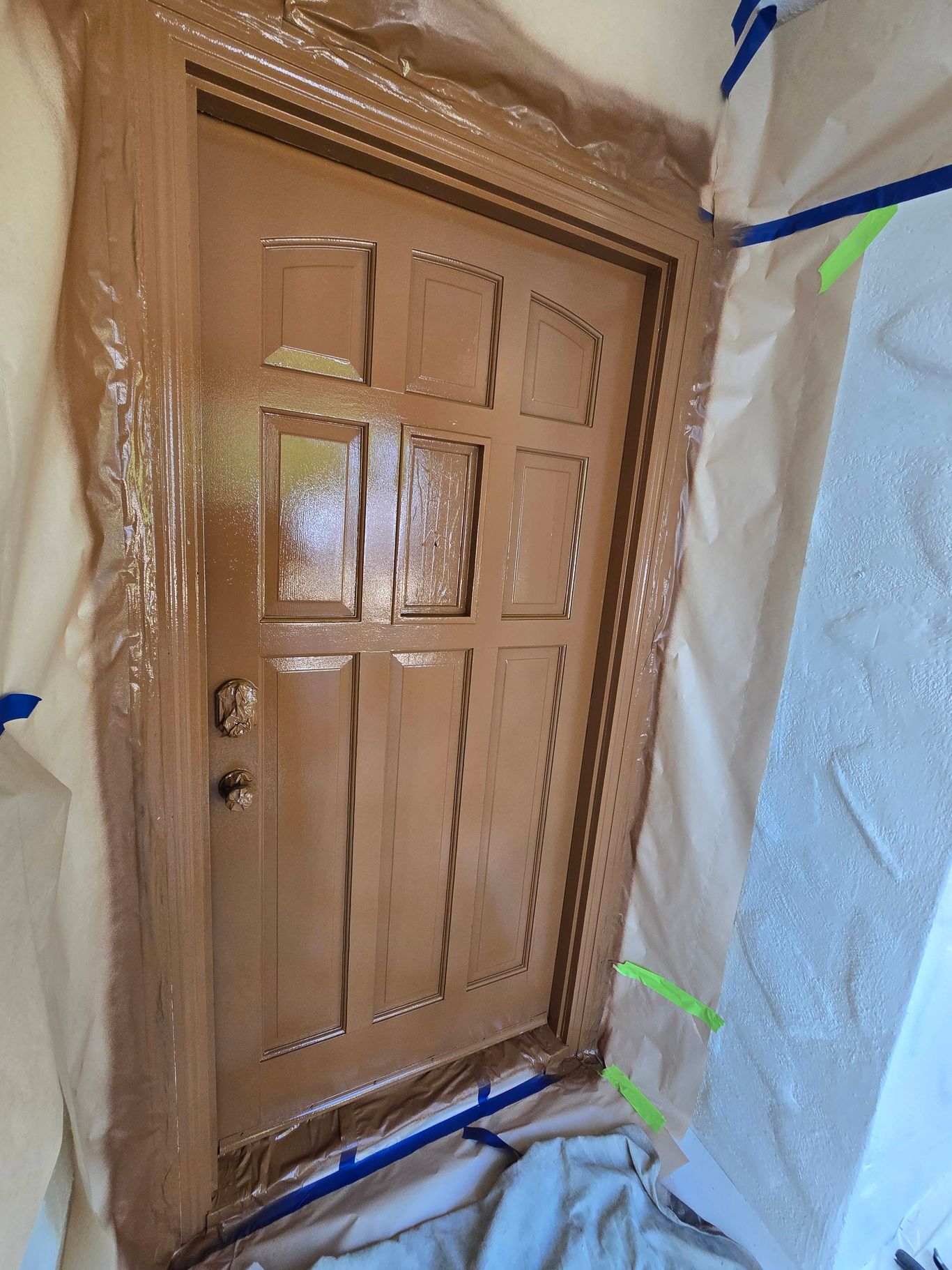 A brown, six-panel door being painted, surrounded by protective plastic sheeting and blue painter's tape.
