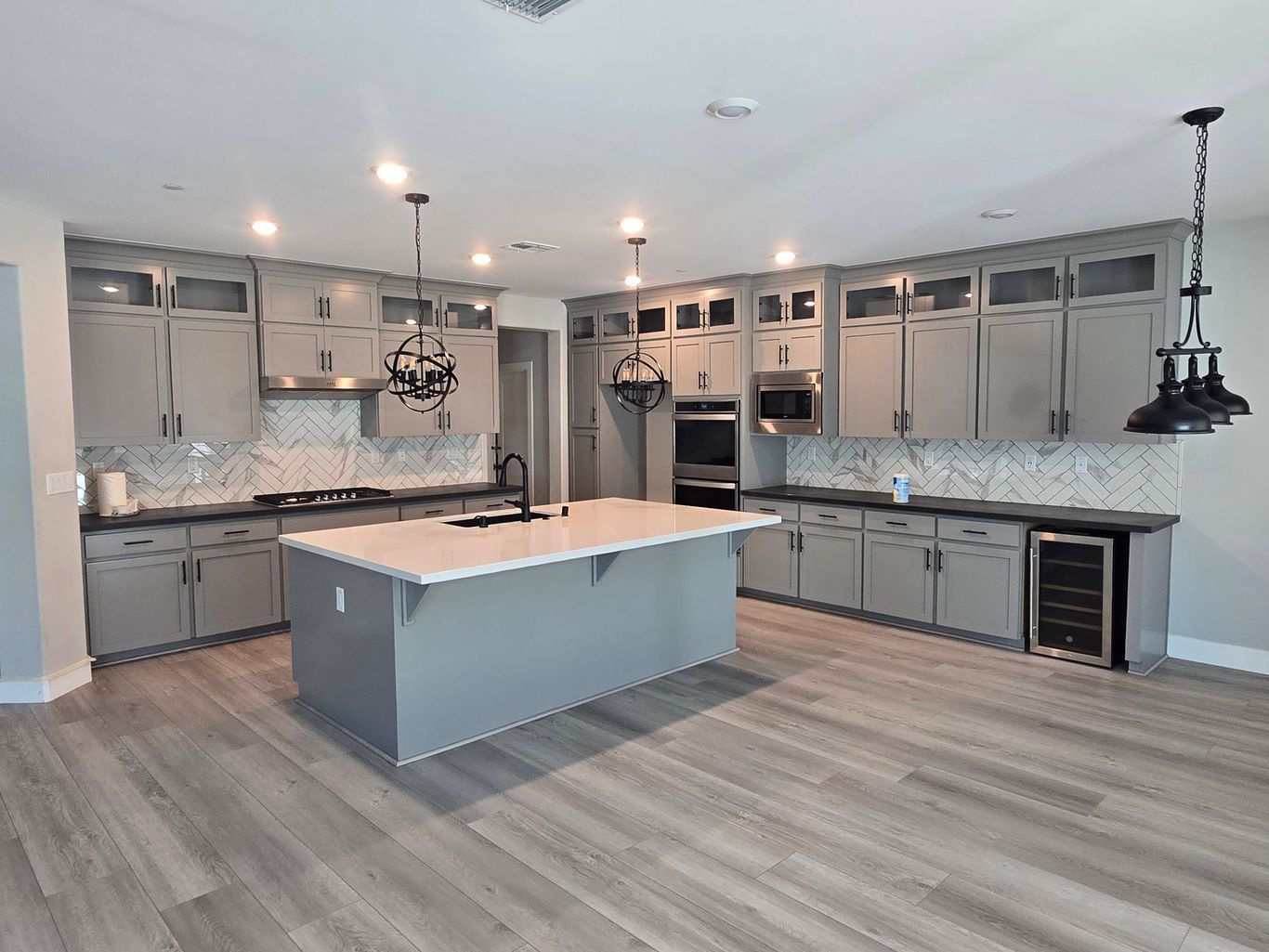 A modern kitchen with grey cabinets, a large central island, white marble-style backsplash, and dark pendant lighting.