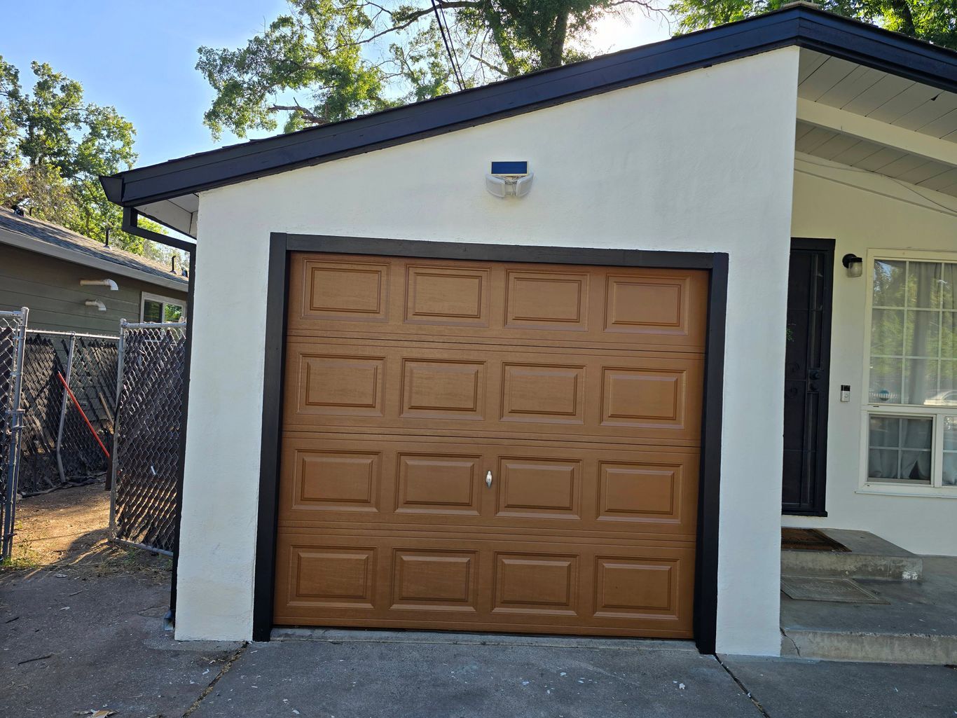 A white garage with a brown, paneled door and black trim, set against a backdrop of trees and a neighboring fence.