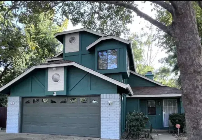A two-story house with dark teal siding, white trim, and a grey garage door, surrounded by trees.