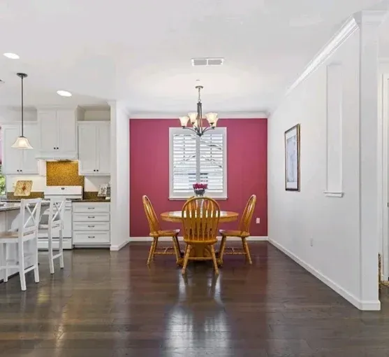 Dining room with a round wooden table and four chairs against a fuchsia accent wall, adjacent to a white kitchen.