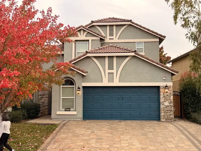 A two-story grey stucco house with a blue garage door, tiled roof, stone accents, and a red-leafed tree in the front yard.