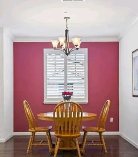 A dining room with a round wooden table, three chairs, and a chandelier centered in front of a berry-colored accent wall.