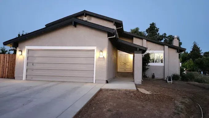 A beige, single-story house with a white double-car garage and a gated front entry at dusk.