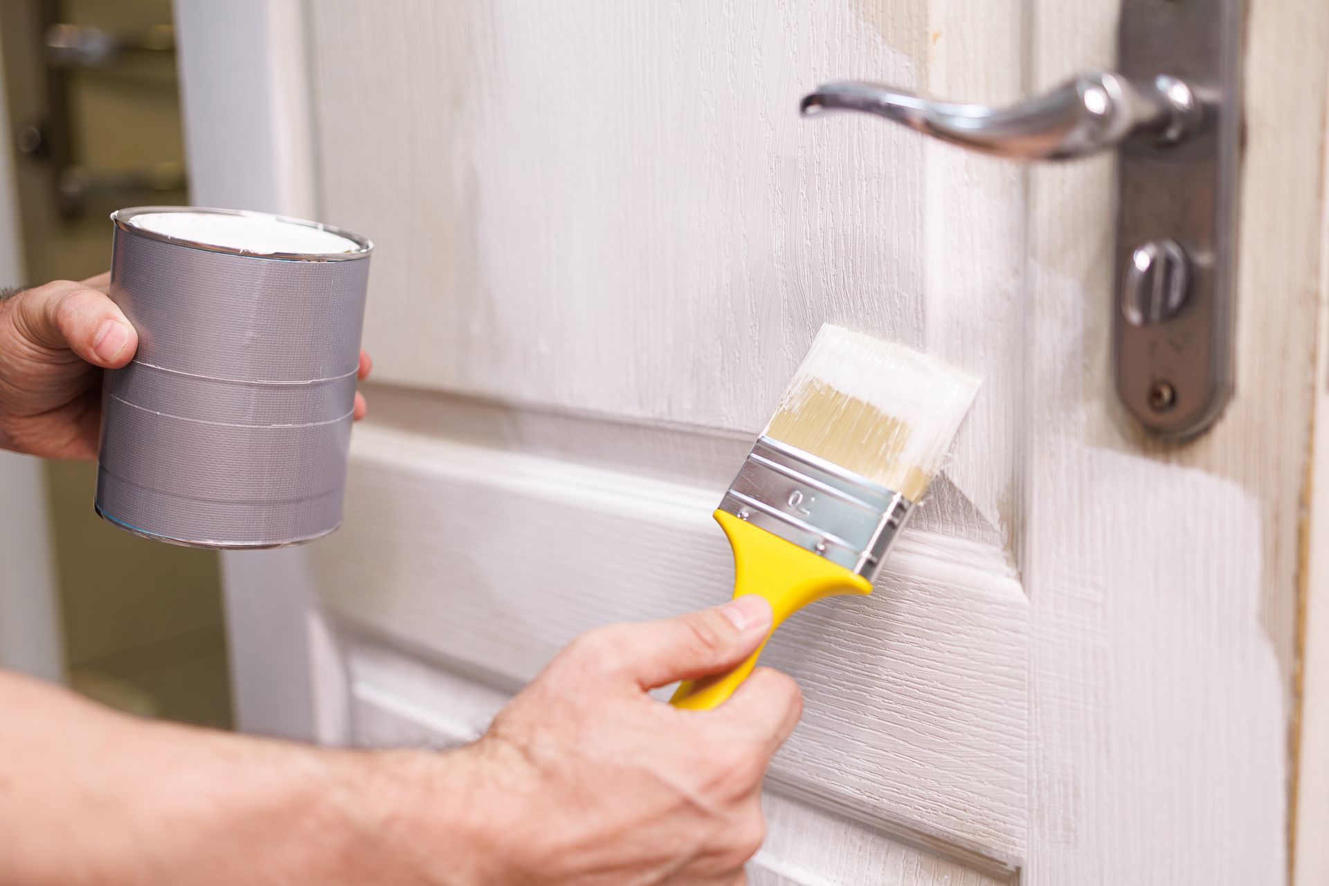 A hand holding a yellow-handled brush paints a white door near a metal handle while holding a paint container.