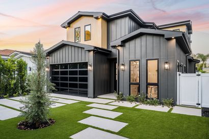 A two-story, modern dark grey home with vertical paneling, a glass garage door, and a grass lawn with concrete pavers.