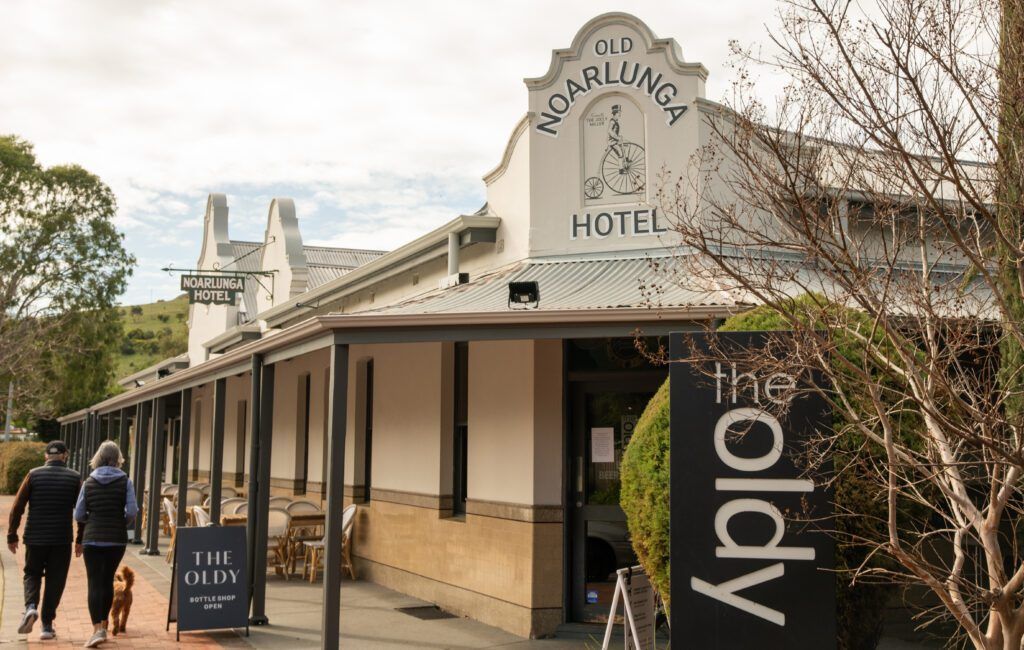 The Old Noarlunga Hotel facade with sign, two people walking in front, cloudy sky.